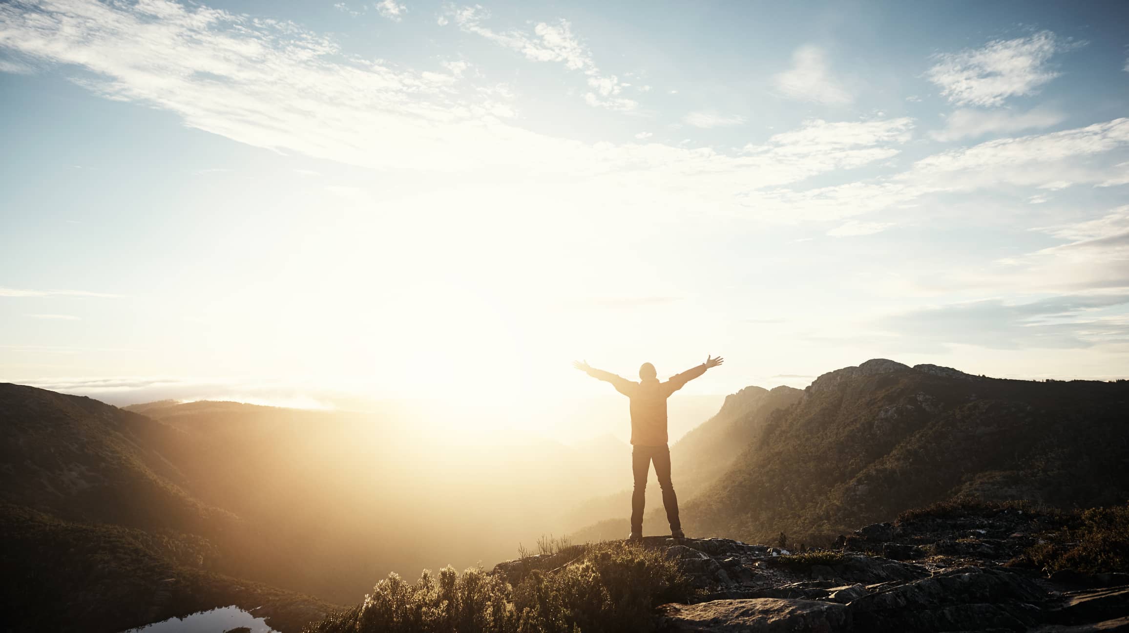 Man standing on a mountain with his arms stretched out while looking at the sun in the distance
