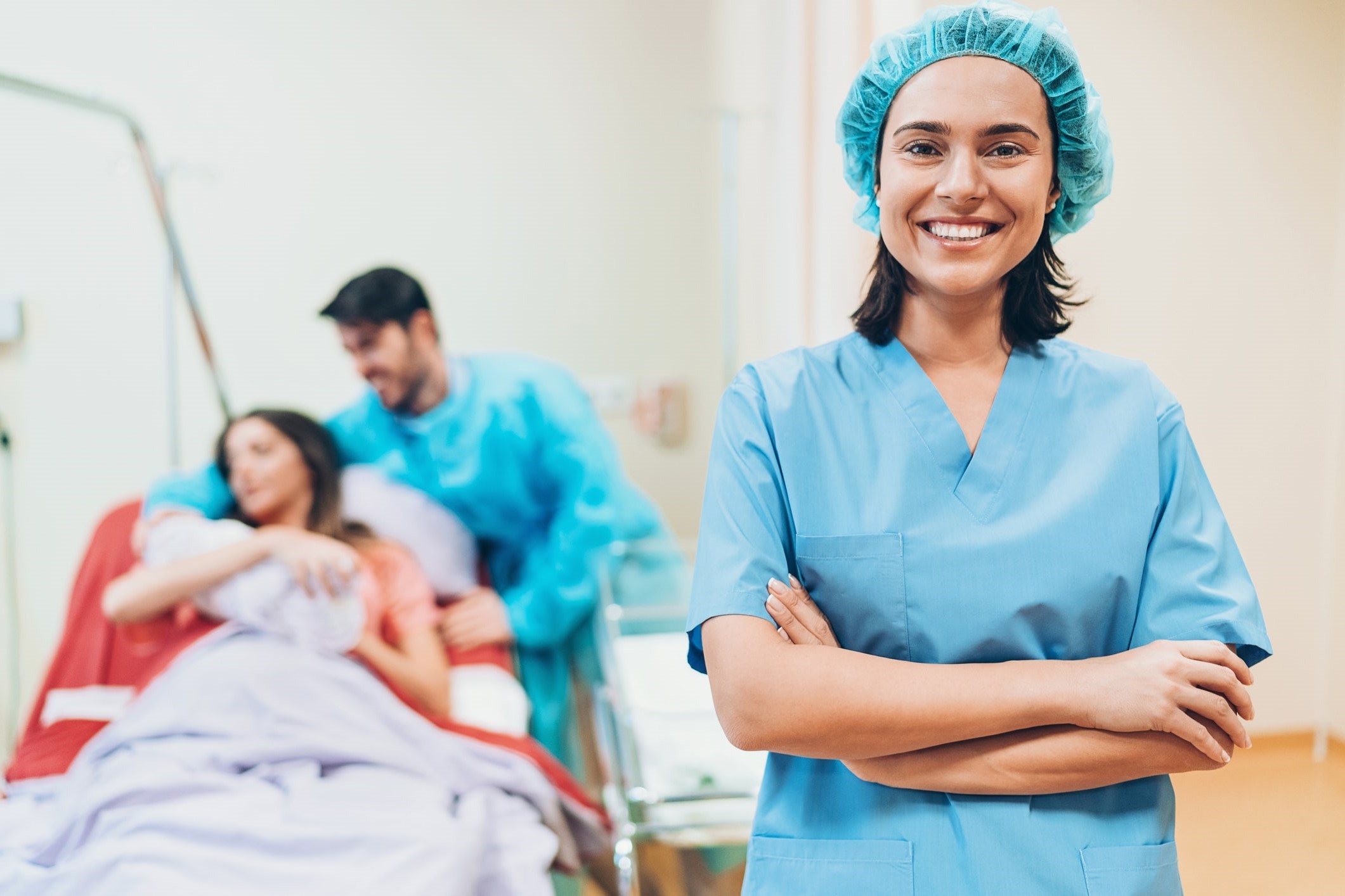 smiling labor and delivery travel nurse stands in front of new parents and baby 