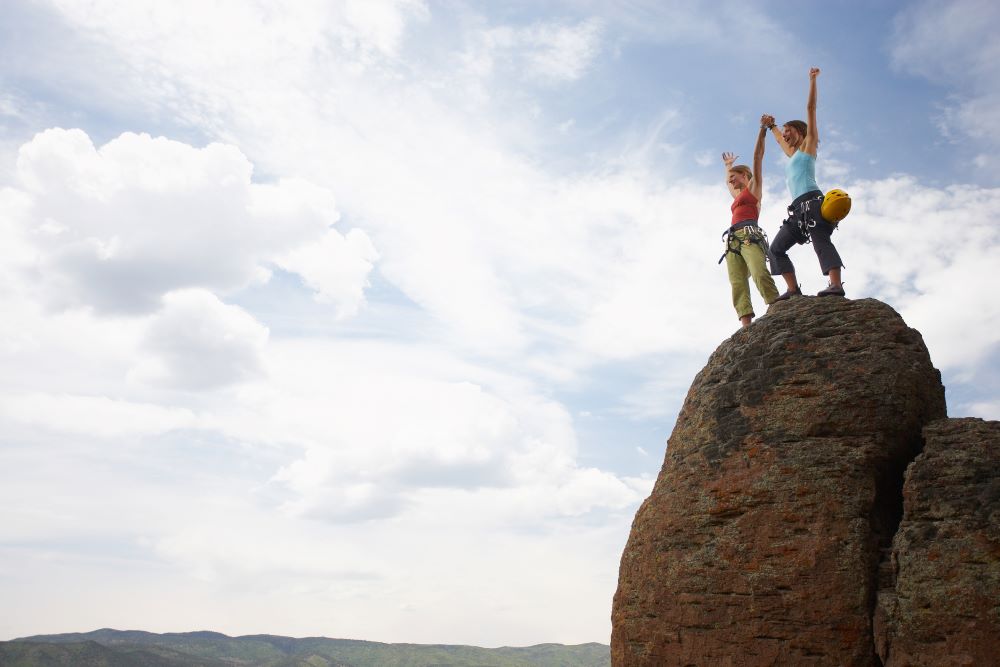 Two female hikers standing on a mountain top with arms raised high